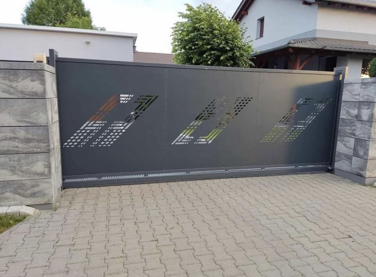 Duża, przesuwna brama wjazdowa w kolorze antracytowym, z pionowymi, betonowymi słupkami po obu stronach. Na bramie znajdują się geometryczne, wycięte wzory przypominające logo i litery z rastrowym efektem, przez które prześwituje światło. Podjazd wyłożony jest szarymi kostkami brukowymi.