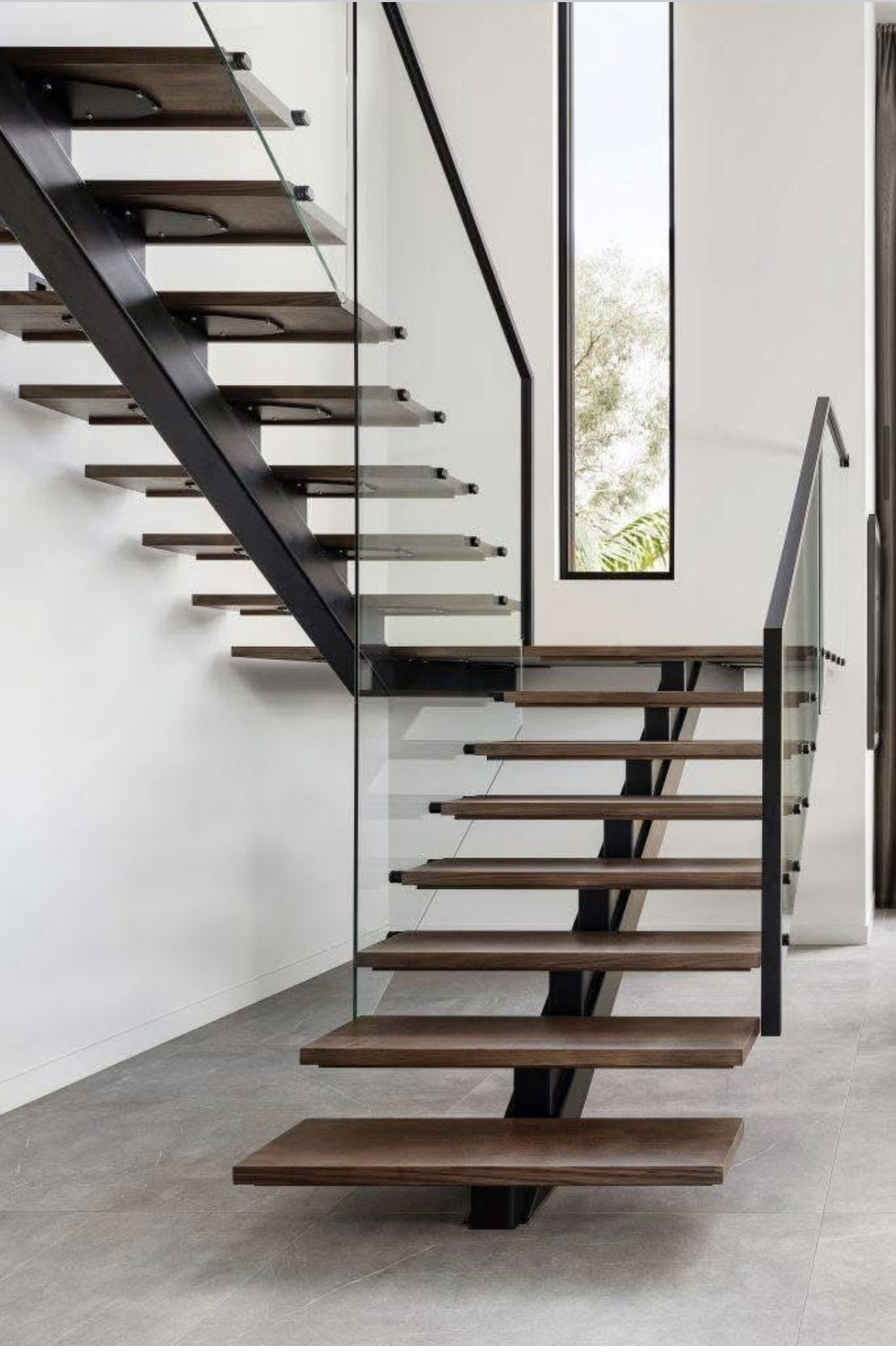 Nowoczesne schody jednobiegowe z metalową konstrukcją nośną w kolorze czarnym, drewnianymi stopniami w jasnym odcieniu oraz elegancką balustradą ze szkła hartowanego i czarnego, minimalistycznego pochwytu. W tle biała ściana i wysokie, pionowe okno.